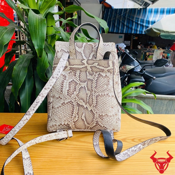 Phụ Kiện Thời Trang Tiện Ích Với Balo Da Trăn Đầy Cá Tính Balo da trăn màu be họa tiết trăn đặt trên bàn gỗ, nền lá xanh và khung cảnh ngoài trời, logo đỏ góc phải.
