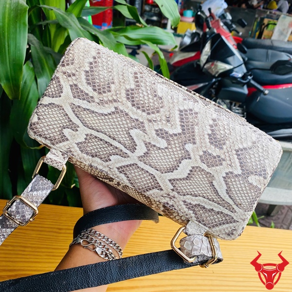 Tỏa Sáng Với Balo Da Trăn Đầy Cá Tính Thời Thượng Tiện Lợi Balo da trăn màu be họa tiết trăn đặt trên bàn gỗ, nền lá xanh và khung cảnh ngoài trời, logo đỏ góc phải.