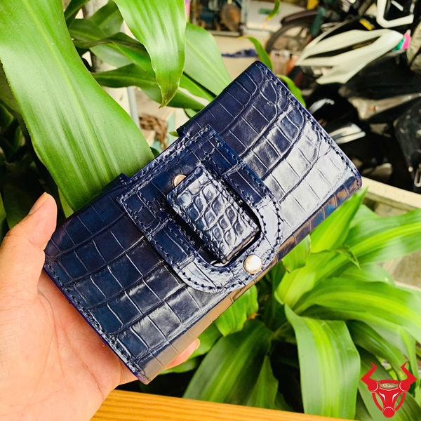 Bao Da Đeo Thắt Lưng Da Cá Sấu Xanh Navy OFAA8 – Cao Cấp Quý Ông Bao da đeo thắt lưng da bụng cá sấu màu xanh navy OFAA8, cầm trên tay, nền lá xanh, logo đỏ góc phải.