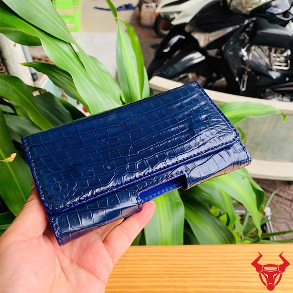 Bao Da Đeo Thắt Lưng Da Bụng Cá Sấu Xanh Navy OFAA8 – Đẳng Cấp Sang Trọng Bao da đeo thắt lưng da bụng cá sấu màu xanh navy OFAA8, cầm trên tay, nền lá xanh, logo đỏ góc phải.