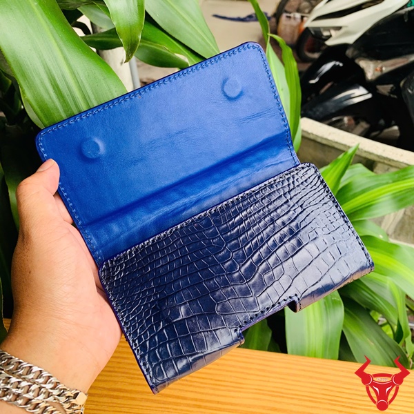 Bao Da Đeo Thắt Lưng Da Cá Sấu Xanh Navy OFAA8 – Sang Trọng Cao Cấp Quý Ông Bao da đeo thắt lưng da bụng cá sấu màu xanh navy OFAA8, cầm trên tay, nền lá xanh, logo đỏ góc phải.
