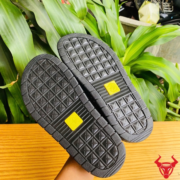 Tự Tin Sải Bước Với Dép Da Trăn DUA8 Thời Trang Êm Ái Đôi dép 2 quai da trăn DUA8 màu nâu đen trên bàn gỗ, nền lá xanh, logo đỏ góc phải.