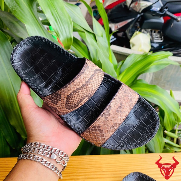 Dép DUA8 Da Trăn – Thời Trang Bền Bỉ, Êm Ái Tuyệt Đối Đôi dép 2 quai da trăn DUA8 màu nâu đen trên bàn gỗ, nền lá xanh, logo đỏ góc phải.