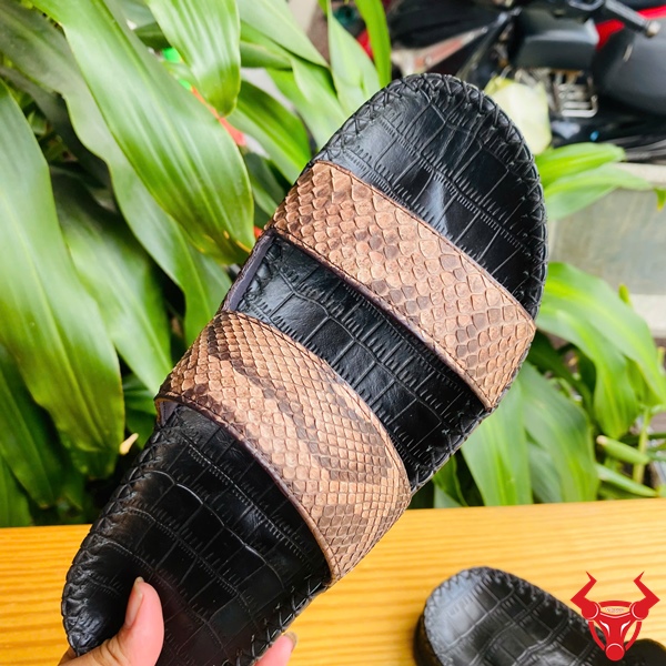 Nâng Tầm Phong Cách Với Dép DUA8 Da Trăn Êm Ái Đôi dép 2 quai da trăn DUA8 màu nâu đen trên bàn gỗ, nền lá xanh, logo đỏ góc phải.
