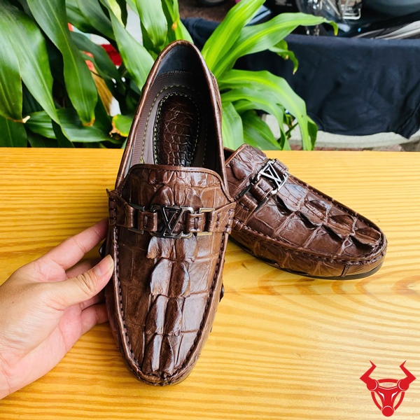 Giày Lười GCA2 – Phong Cách Đẳng Cấp Sang Trọng Và Êm Ái Đôi giày lười da đuôi cá sấu GCA2 màu nâu, khóa LV, đặt trên bàn gỗ, nền lá xanh, logo đỏ góc phải.