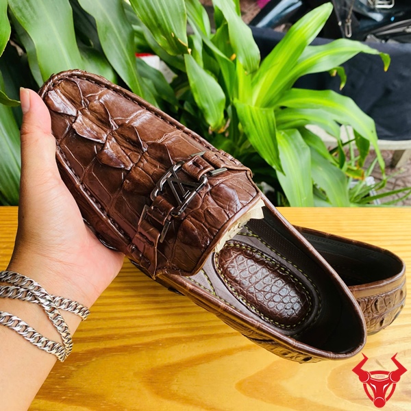 Giày Lười GCA2 Da Cá Sấu – Thời Trang Sang Trọng Đẳng Cấp Êm Ái Đôi giày lười da đuôi cá sấu GCA2 màu nâu, khóa LV, đặt trên bàn gỗ, nền lá xanh, logo đỏ góc phải.