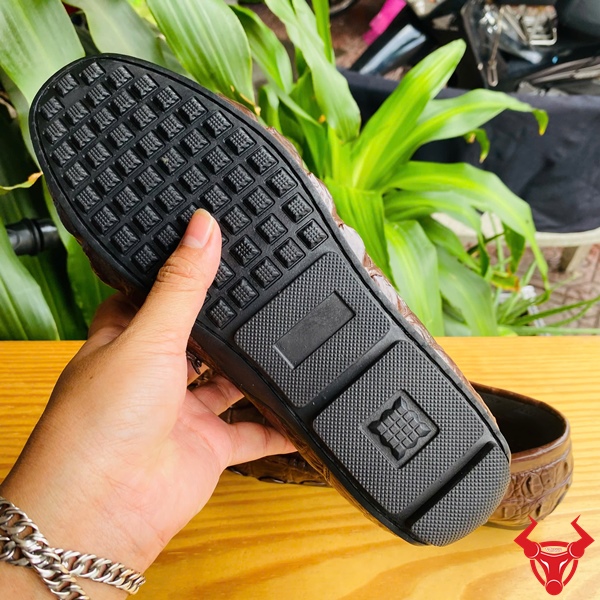 Phong Cách Đẳng Cấp Với Giày Lười GCA2 Da Cá Sấu Sang Trọng Êm Ái Đôi giày lười da đuôi cá sấu GCA2 màu nâu, khóa LV, đặt trên bàn gỗ, nền lá xanh, logo đỏ góc phải.