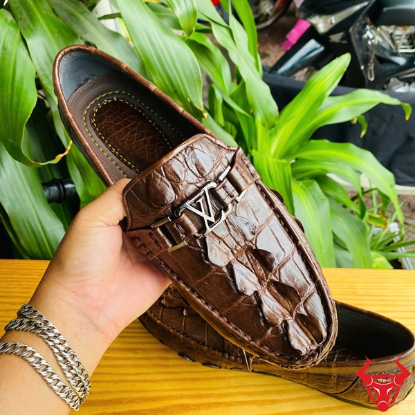Giày Lười Da Cá Sấu GCA2 – Thời Trang Sang Trọng Êm Ái Đẳng Cấp Đôi giày lười da đuôi cá sấu GCA2 màu nâu, khóa LV, đặt trên bàn gỗ, nền lá xanh, logo đỏ góc phải.