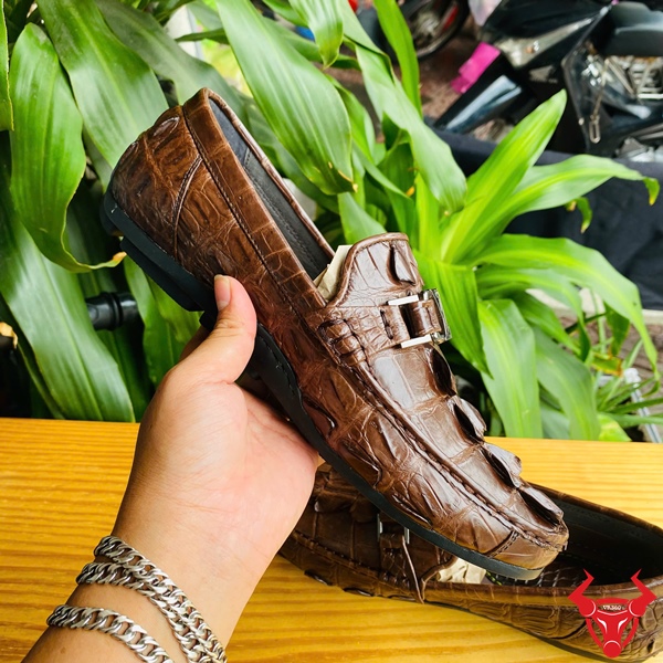 Nâng Tầm Phong Cách Với Giày Lười GCA2 Sang Trọng Đẳng Cấp Êm Ái Đôi giày lười da đuôi cá sấu GCA2 màu nâu, khóa LV, đặt trên bàn gỗ, nền lá xanh, logo đỏ góc phải.