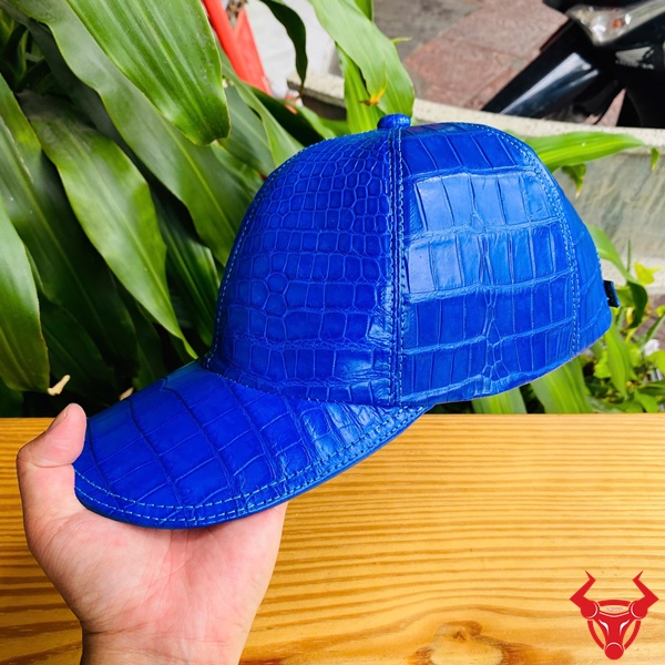 NOA8 Xanh Dương Phong Cách Cá Tính Đẳng Cấp Mũ lưỡi trai xanh dương da bụng cá sấu trên bàn gỗ, tay người cầm, nền lá xanh tươi.
