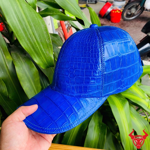 Sắc Xanh Dương Sang Trọng Của Mũ NOA8 Mũ lưỡi trai xanh dương từ da cá sấu, trên bàn gỗ, tay cầm, nền lá xanh.