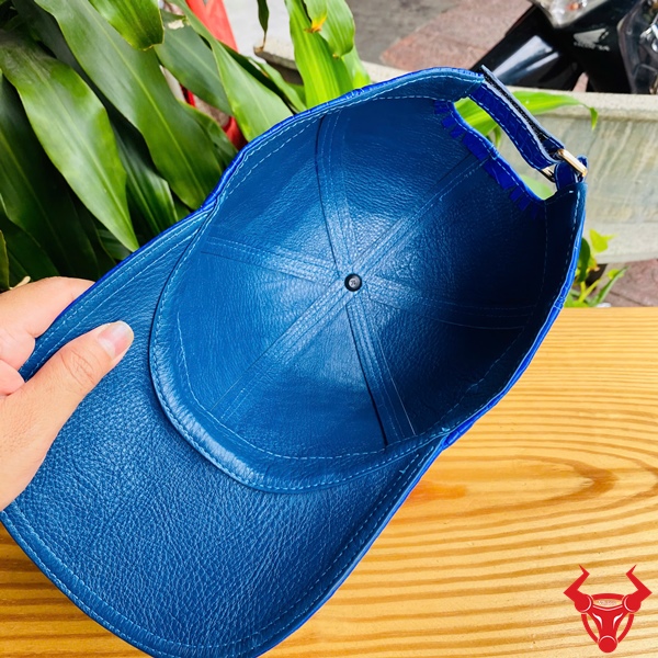 Mũ Lưỡi Trai NOA8 Xanh Dương Quyến Rũ Sang Trọng Mũ lưỡi trai xanh dương từ da cá sấu trên bàn gỗ, tay cầm, nền lá xanh tươi.
