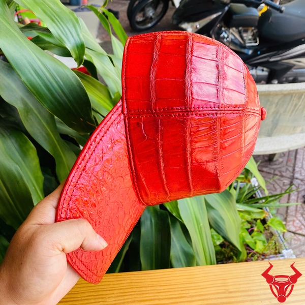 Mũ Lưỡi Trai Da Bụng Cá Sấu NOA8 – Cá Tính Cao Cấp Thời Thượng Mũ lưỡi trai da bụng cá sấu NOA8 màu đỏ, đặt trên bàn gỗ, nền lá xanh, logo đỏ góc phải.