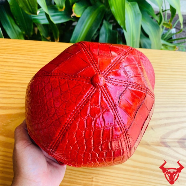 Mũ Lưỡi Trai Da Cá Sấu NOA8 – Thời Thượng Đẳng Cấp Cá Tính Mũ lưỡi trai da bụng cá sấu NOA8 màu đỏ, đặt trên bàn gỗ, nền lá xanh, logo đỏ góc phải.