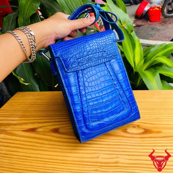 Túi Xanh Dương Đậm Màu Sắc Cá Nhân Túi đeo chéo xanh dương da cá sấu trên bàn gỗ, dây đeo, nền lá xanh và xe máy.