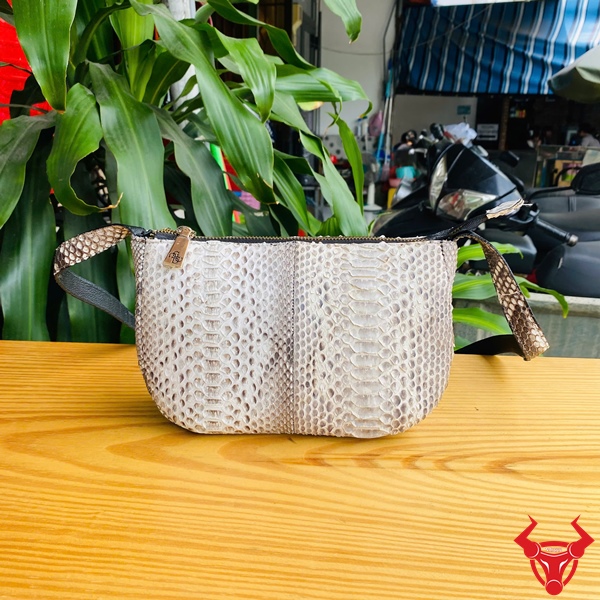 Phụ Kiện Thời Trang Cá Tính Với Túi Đeo Nữ Da Trăn Tiện Lợi Túi đeo nữ da trăn màu trắng họa tiết trăn đặt trên bàn gỗ, dây đeo đen, nền lá xanh và khung cảnh ngoài trời, logo đỏ góc phải.