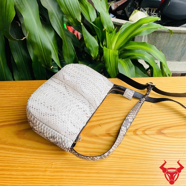 Túi Đeo Nữ Da Trăn – Thời Trang Cá Tính Tiện Lợi Hiện Đại Túi đeo nữ da trăn màu trắng họa tiết trăn đặt trên bàn gỗ, dây đeo đen, nền lá xanh và khung cảnh ngoài trời, logo đỏ góc phải.