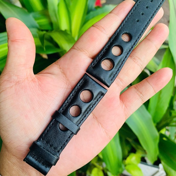 Dây đồng hồ da bò thời trang độc đáo Hình ảnh dây đồng hồ da bò đen, thiết kế riêng, thẩm mỹ, cầm trên tay với cây xanh phía sau.