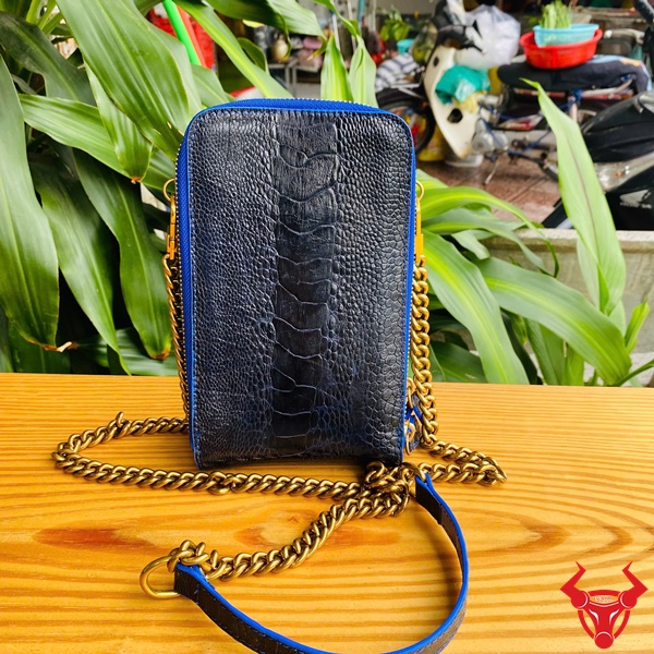 Túi Da Chân Đà Điểu - Tiện Dụng Và Sang Trọng Hình ảnh túi đeo chéo da đà điểu xanh navy với dây xích vàng, trên bàn gỗ, nền cây xanh, nhấn mạnh sự cao cấp và tiện lợi.