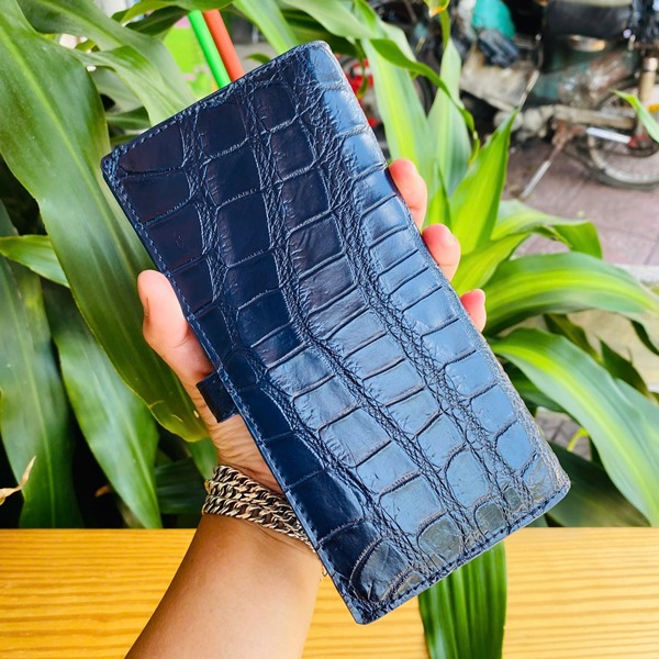 Ví da cá sấu Thái Lan sang trọng vượt trội Hình ảnh ví da cá sấu Thái Lan hai gấp, xanh đậm, khóa kim loại, quý phái, trên bàn gỗ với cây xanh.