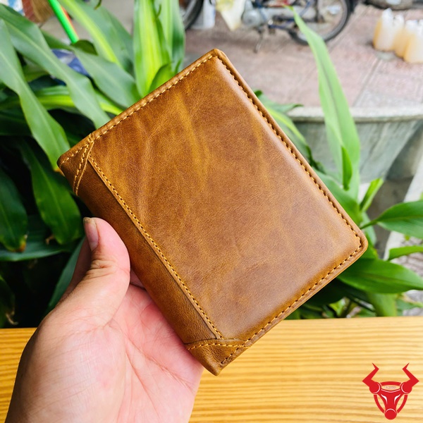 Ví Da Bò Màu Nâu Bò - Phong Cách Tinh Tế Hình ảnh ví da bò nâu nhiều ngăn, cầm tay, nền cây xanh, nhấn mạnh sự tiện lợi và sang trọng.