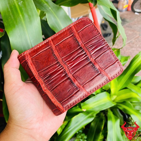 Ví VBA8 Nâu Đỏ Cao Cấp Trong Thời Trang Hiện Đại Ví ngang da cá sấu màu nâu đỏ VBA8, tay cầm, nền cây xanh và xe máy.