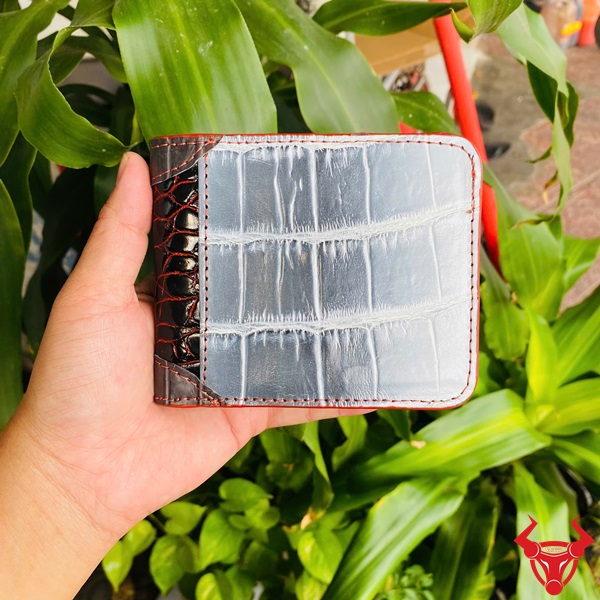 Ví Ngang Da Cá Sấu Phối Màu Thời Trang Độc Lạ Ví ngang da cá sấu phối màu trắng và nâu, tay cầm, nền lá xanh và xe máy phía sau.