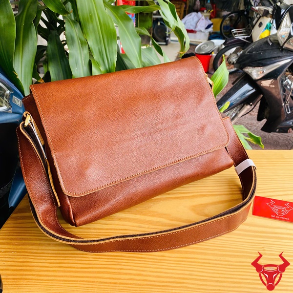 Túi Đeo Chéo Da Bò TN06 - Thời Trang Năng Động Hình ảnh túi đeo chéo da bò form ngang TN06 màu nâu, phong cách năng động, đặt trên bàn gỗ với nền cây xanh và xe máy.