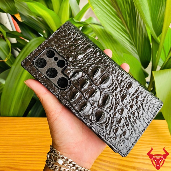 Phụ Kiện Chắc Chắn Sang Trọng Bền Bỉ Cao Cấp Đẳng Cấp Case S24 Ultra Da Cá Sấu Gù Lưng Màu Đen Ảnh close-up bao da S24 Ultra da cá sấu gù lưng đen, cầm tay với logo, nền xanh, nhấn mạnh phụ kiện cao cấp.