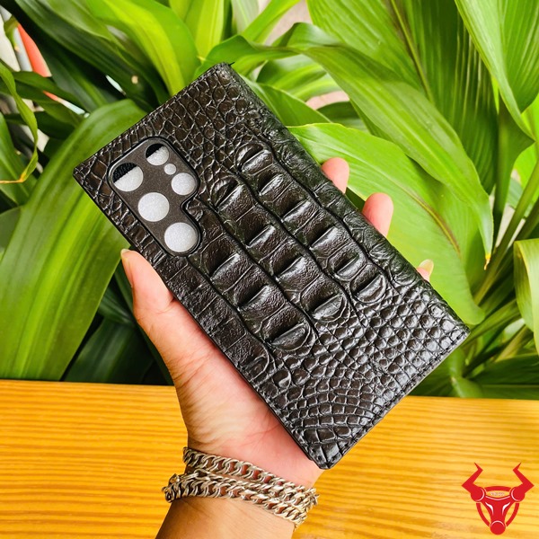 Phụ Kiện Chắc Chắn Sang Trọng Bền Bỉ Cao Cấp Đẳng Cấp Case S24 Ultra Da Cá Sấu Màu Đen Ảnh close-up bao da S24 Ultra da cá sấu đen, cầm tay với logo, nền xanh, nhấn mạnh phụ kiện cao cấp.