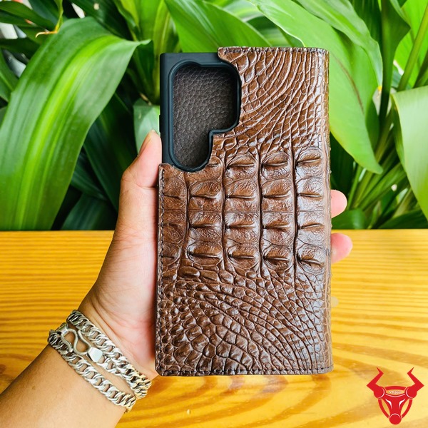 Phụ Kiện Cao Cấp Sang Trọng Bền Bỉ Chắc Chắn Đẳng Cấp Bao Da Samsung S24 Ultra Da Cá Sấu Gai Lưng Nâu Đất Hình ảnh bao da Samsung S24 Ultra da cá sấu gai lưng nâu đất, cầm tay trước nền lá xanh, thể hiện sự cao cấp và chắc chắn.