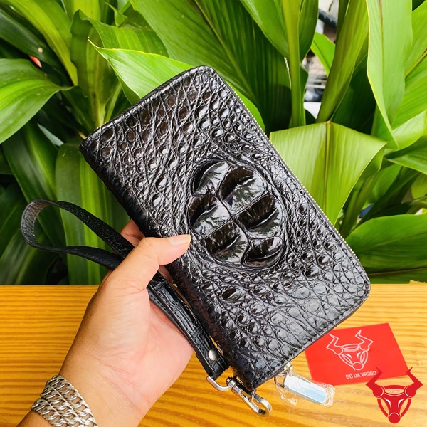 Đẳng Cấp Sang Trọng: Ví Da Cá Sấu BUA4 Đen Thời Trang Cao Cấp Bền Bỉ Ảnh ví da cá sấu đen cầm tay, nền thực vật xanh, kèm thẻ thương hiệu đỏ.