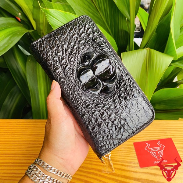 Thời Trang Sang Trọng: Ví Cầm Tay BUA4 Đen Đẳng Cấp Cao Cấp Bền Bỉ Hình người cầm ví da cá sấu đen sang trọng, nền lá xanh, thẻ thương hiệu bên cạnh.