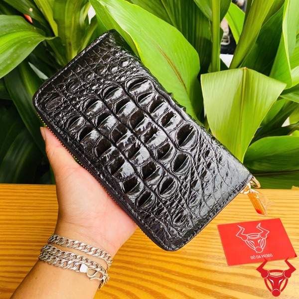 Cao Cấp Đẳng Cấp: Ví Da Cá Sấu Gù BUA4 Đen Thời Trang Sang Trọng Bền Bỉ Ảnh ví da cá sấu đen cao cấp cầm tay, nền lá xanh, kèm thẻ thương hiệu đỏ.