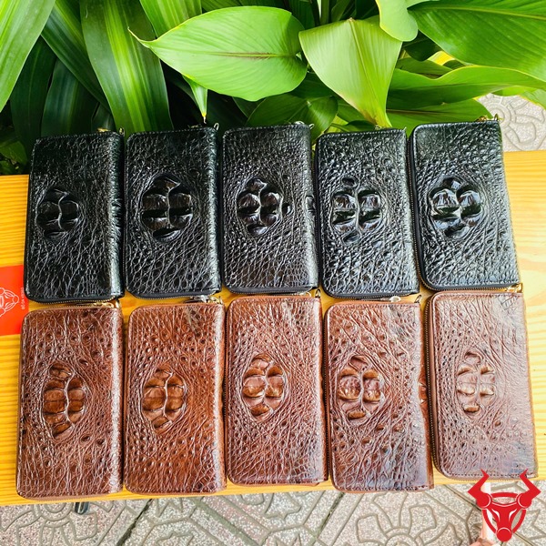 Sang Trọng Cao Cấp: Ví BUA4 Đen Thời Trang Đẳng Cấp Bền Bỉ Ảnh ví da cá sấu đen cao cấp cầm tay, nền lá xanh, thẻ thương hiệu đỏ bên cạnh.