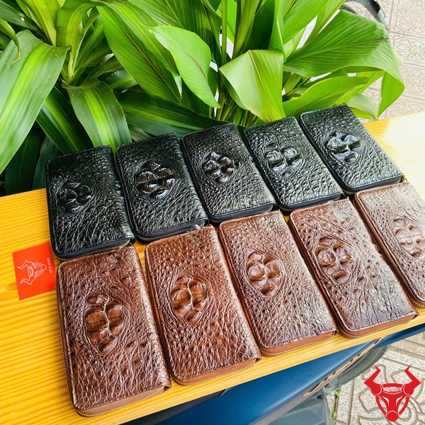 Bền Bỉ Cao Cấp: Ví Cầm Tay Gù BUA4 Đen Thời Trang Sang Trọng Đẳng Cấp Hình ảnh ví da cá sấu đen cao cấp được cầm, nền lá xanh, kèm thẻ thương hiệu đỏ.