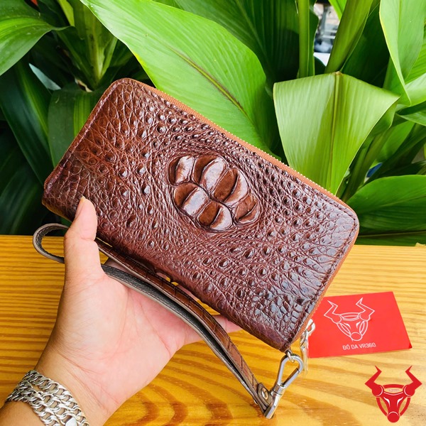 Đẳng Cấp Sang Trọng: Ví Da Cá Sấu BUA4 Nâu Đất Thời Trang Cao Cấp Bền Bỉ Ảnh ví da cá sấu nâu đất cầm tay, nền thực vật xanh, kèm thẻ thương hiệu đỏ.
