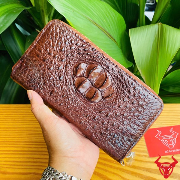 Thời Trang Sang Trọng: Ví Cầm Tay BUA4 Nâu Đất Đẳng Cấp Cao Cấp Bền Bỉ Hình người cầm ví da cá sấu nâu đất sang trọng, nền lá xanh, thẻ thương hiệu bên cạnh.