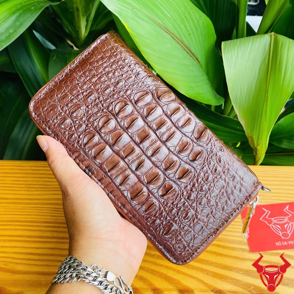 Cao Cấp Đẳng Cấp: Ví Da Cá Sấu Gù BUA4 Nâu Đất Thời Trang Sang Trọng Bền Bỉ Ảnh ví da cá sấu nâu đất cao cấp cầm tay, nền lá xanh, kèm thẻ thương hiệu đỏ.