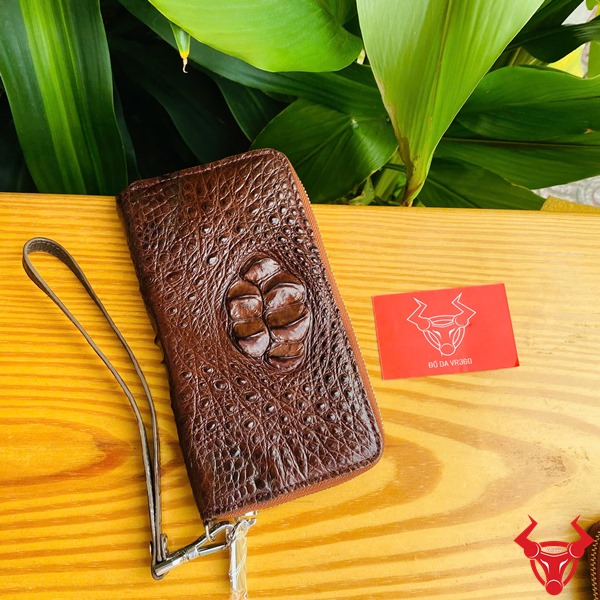 Sang Trọng Thời Trang: Ví Nam Nữ BUA4 Nâu Đất Đẳng Cấp Cao Cấp Bền Bỉ Hình ảnh ví da cá sấu nâu đất cầm tay, nền lá xanh, thẻ thương hiệu đỏ bên cạnh.