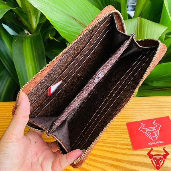 Đẳng Cấp Cao Cấp: Ví Cầm Tay Da Cá Sấu BUA4 Nâu Đất Sang Trọng Bền Bỉ Thời Trang Ảnh người cầm ví da cá sấu nâu đất cao cấp, nền lá xanh, thẻ thương hiệu bên cạnh.