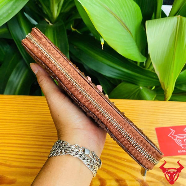Thời Trang Đẳng Cấp: Ví Da Gù BUA4 Nâu Đất Sang Trọng Cao Cấp Bền Bỉ Hình ví da cá sấu nâu đất sang trọng cầm tay, nền lá xanh, thẻ thương hiệu đỏ.