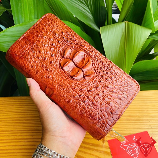 Đẳng Cấp Sang Trọng: Ví Da Cá Sấu BIA4 Nâu Đỏ Thời Trang Cao Cấp Tiện Dụng Ảnh ví da cá sấu nâu đỏ cầm tay, nền thực vật xanh, kèm thẻ thương hiệu đỏ.