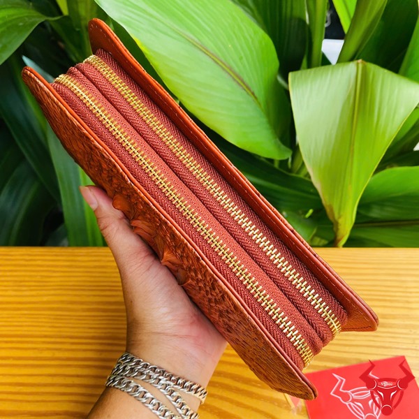 Thời Trang Sang Trọng: Ví Cầm Tay BIA4 Nâu Đỏ Đẳng Cấp Cao Cấp Tiện Lợi Hình người cầm ví da cá sấu nâu đỏ sang trọng, nền lá xanh, thẻ thương hiệu bên cạnh.