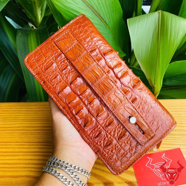 Cao Cấp Đẳng Cấp: Ví Da Cá Sấu Gù BIA4 Nâu Đỏ Thời Trang Sang Trọng Tiện Dụng Ảnh ví da cá sấu nâu đỏ cao cấp, nền lá xanh, kèm thẻ thương hiệu đỏ.