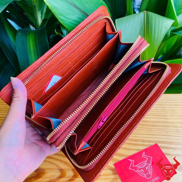 Sang Trọng Thời Trang: Ví Nam Nữ BIA4 Nâu Đỏ Đẳng Cấp Cao Cấp Tiện Dụng Hình ảnh ví da cá sấu nâu đỏ cầm tay, nền lá xanh, thẻ thương hiệu đỏ bên cạnh.