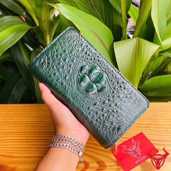 Đẳng Cấp Sang Trọng: Ví Da Cá Sấu BIA4 Xanh Lá Thời Trang Cao Cấp Tiện Dụng Ảnh ví da cá sấu xanh lá cầm tay, nền thực vật xanh, kèm thẻ thương hiệu đỏ.