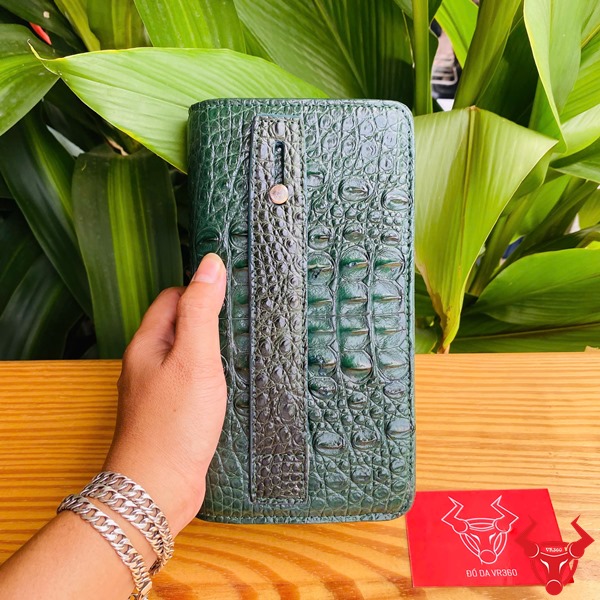 Thời Trang Sang Trọng: Ví Cầm Tay BIA4 Xanh Lá Đẳng Cấp Cao Cấp Tiện Lợi Hình người cầm ví da cá sấu xanh lá sang trọng, nền lá xanh, thẻ thương hiệu bên cạnh.