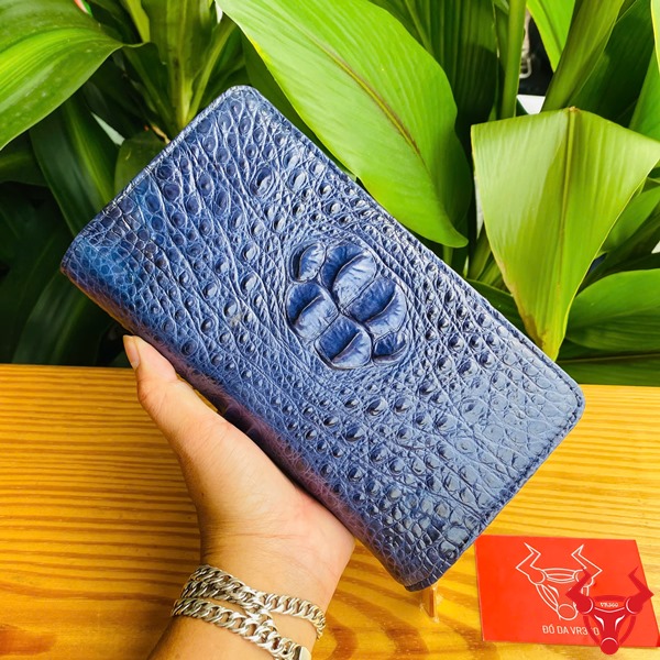Đẳng Cấp Sang Trọng: Ví Da Cá Sấu BIA4 Xanh Navy Thời Trang Cao Cấp Tiện Dụng Ảnh ví da cá sấu xanh navy cầm tay, nền thực vật xanh, kèm thẻ thương hiệu đỏ.