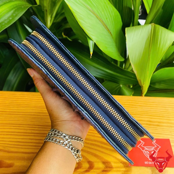 Thời Trang Sang Trọng: Ví Cầm Tay BIA4 Xanh Navy Đẳng Cấp Cao Cấp Tiện Lợi Hình người cầm ví da cá sấu xanh navy sang trọng, nền lá xanh, thẻ thương hiệu bên cạnh.